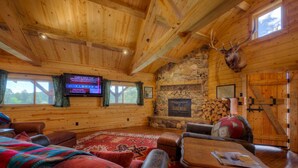 Interior de una cabaña rústica con chimenea de piedra, vigas de madera en el techo, cabeza de venado montada y un televisor mostrando programación deportiva.