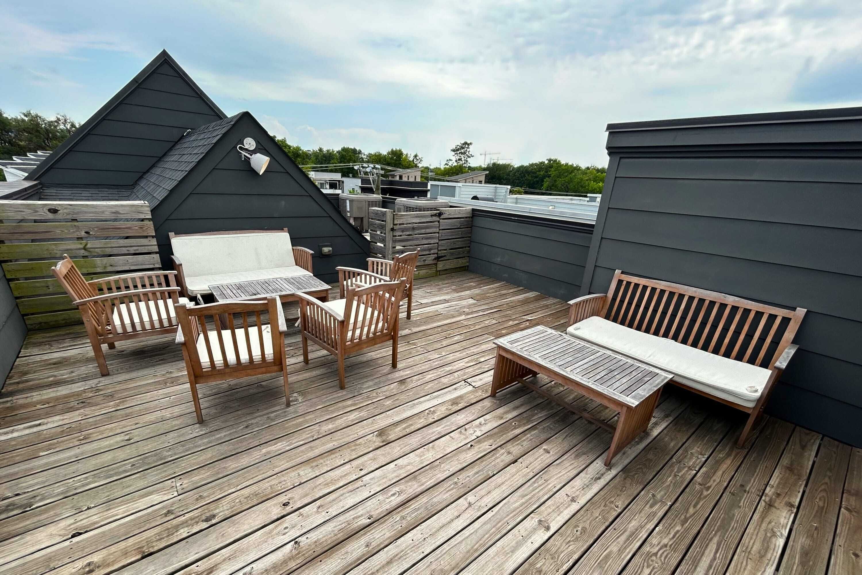 Een houten terras met een tafel, stoelen, een bankje en een zwart gebouw op de achtergrond.