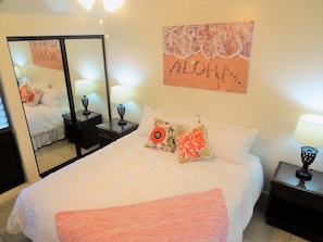 Una cama bien hecha con una manta rosa, un espejo, una mesita de noche con una lámpara y una pared con un letrero que dice 'ALOHA'.