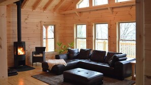 Interior acogedor de una cabaña con estufa de leña, sofá de cuero y ventanas amplias.