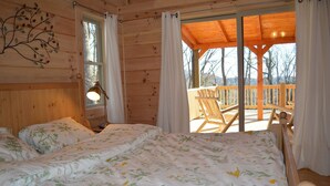 Un dormitorio de cabaña de madera con una cama grande, una mesita de noche, una lámpara y vistas al balcón y al exterior.