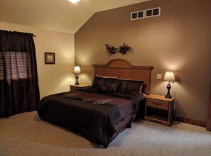 Une chambre avec un grand lit, deux tables de chevet avec des lampes, une tête de lit de couleur sombre, une bouche d’aération fixée au mur et un tableau encadré accroché au mur.