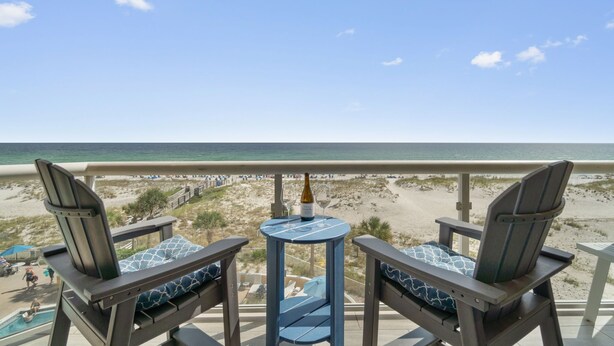 Ein Balkon mit zwei Adirondack-Stühlen, einem kleinen runden Tisch und einer Champagnerflasche. Blick auf Strand und Meer.