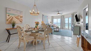 Ein geräumiger Essbereich mit einem runden Tisch, Stühlen und einer Chandelüster. Durch große Fenster ist der Blick aufs Meer möglich.