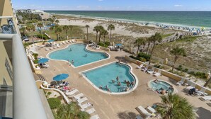 Ein Resort mit zwei Schwimmbecken, umgeben von Liegestühlen und Sonnenschirmen, ein Strand im Hintergrund und Blick aufs Meer.