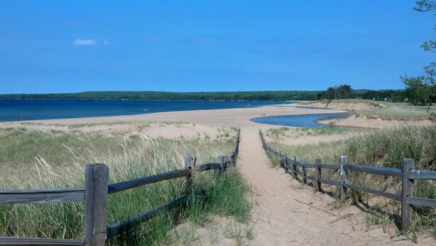 A sandy beach path leading to a body of water, surrounded by grassy dunes and a wooden fence.