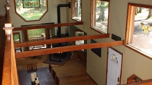A spacious loft-style room with a wooden floor, a dining area with a table and chairs, a large window with a view of trees, and a ceiling fan.