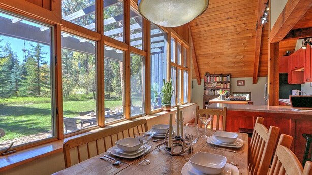 Un comedor con una mesa de madera dispuesta para seis personas, ventanas amplias y vistas a un bosque.