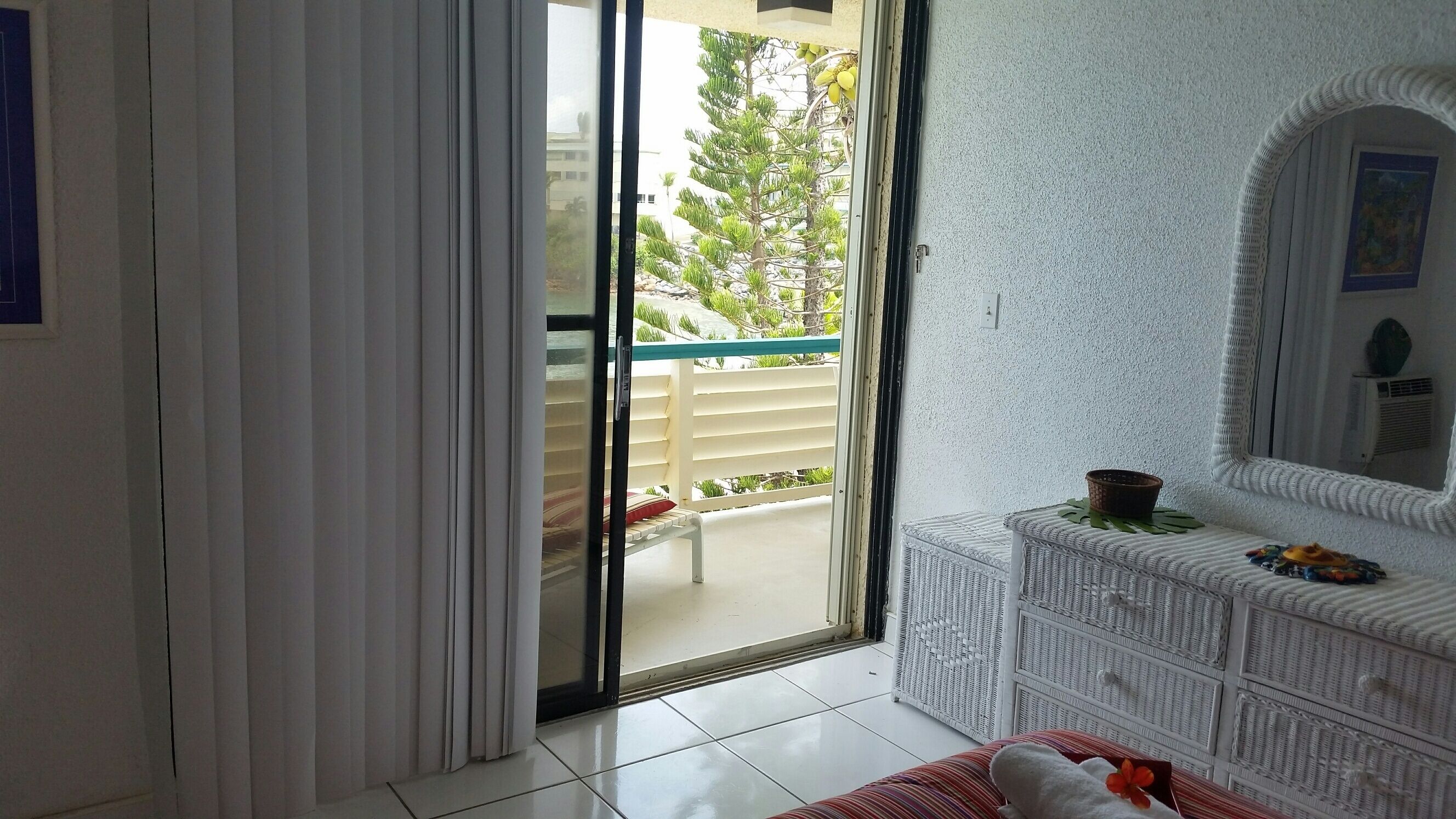 Una habitación con puerta corrediza de vidrio que da a un balcón con barandilla blanca y vistas al verdor.