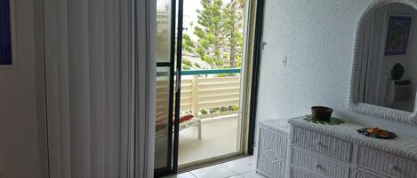 Una habitación con puerta corrediza de vidrio que da a un balcón con barandilla blanca y vistas al verdor.