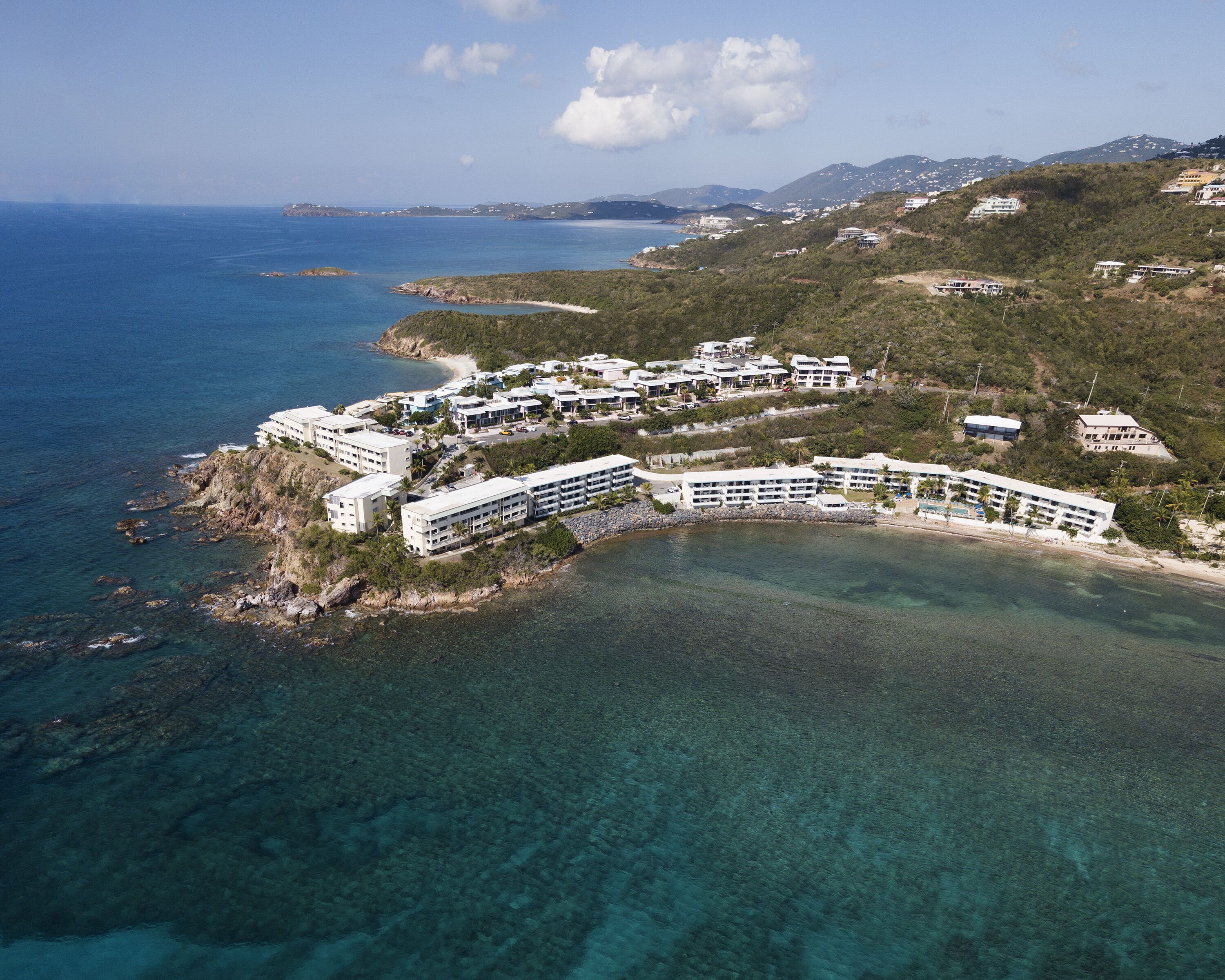 An aerial view of a coastal resort with multiple buildings, a clear blue sea, and a hilly landscape in the background.