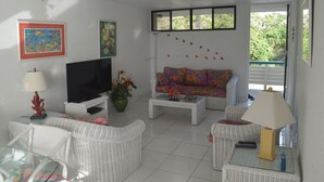 A living room with a TV, white wicker furniture, a colorful sofa, and a window with a view of greenery.