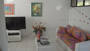 A living room with a flat-screen TV on a white stand, a white wicker coffee table with magazines, a floral sofa, and two wall-mounted paintings.
