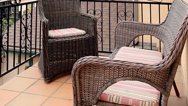 Un balcon avec des fauteuils en osier et une table, donnant sur un quartier résidentiel bordé de palmiers et de maisons.
