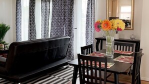 Un salon avec un canapé noir, une table à manger avec des chaises, un vase rempli de fleurs et une fenêtre ornée de rideaux à motifs.