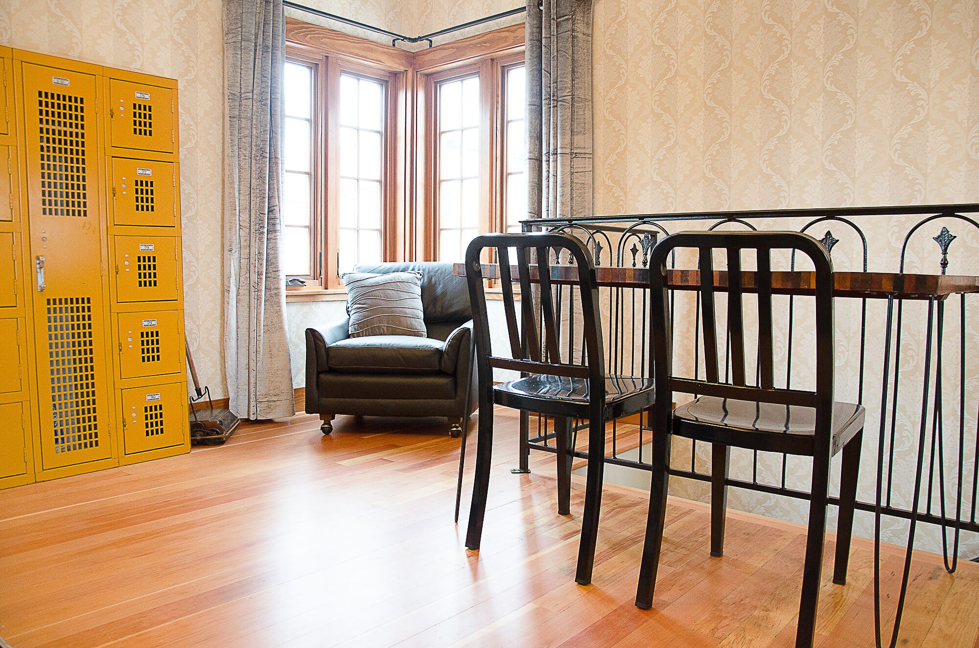 A room with wooden flooring, a black upholstered chair, a yellow metal cabinet, and a window with curtains.