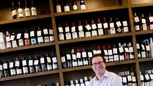 Un hombre sentado en un taburete en una bodega, con estantes llenos de botellas de vino.