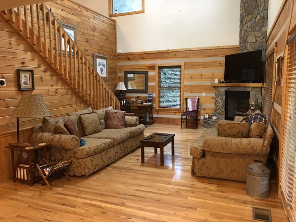A cozy log cabin interior with a wooden staircase, a stone fireplace, and a comfortable seating area with a sofa and armchairs.