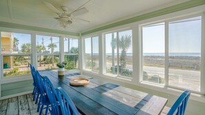 Ruime kamer met een grote tafel en blauwe stoelen, grote ramen met uitzicht op het strand en palmbomen, en een plafondventilator.