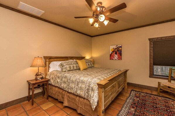 A bedroom with a wooden bed, a nightstand, a lamp, a ceiling fan, a window with blinds, and a patterned rug.