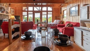 Ein gemütliches Holzblockhaus-Interieur mit einem Esstisch, gedeckt für vier Personen, umgeben von roten Sofas und einem großen Fenster mit schöner Aussicht.