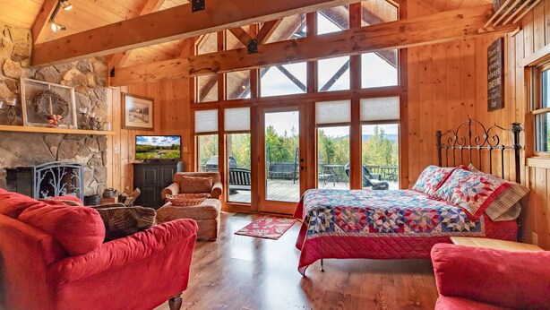 Ein gemütliches Blockhäuschen-Interieur mit Kamin aus Stein, einem großen Bett mit Bettdecke und einem Balkon mit Blick ins Freie.