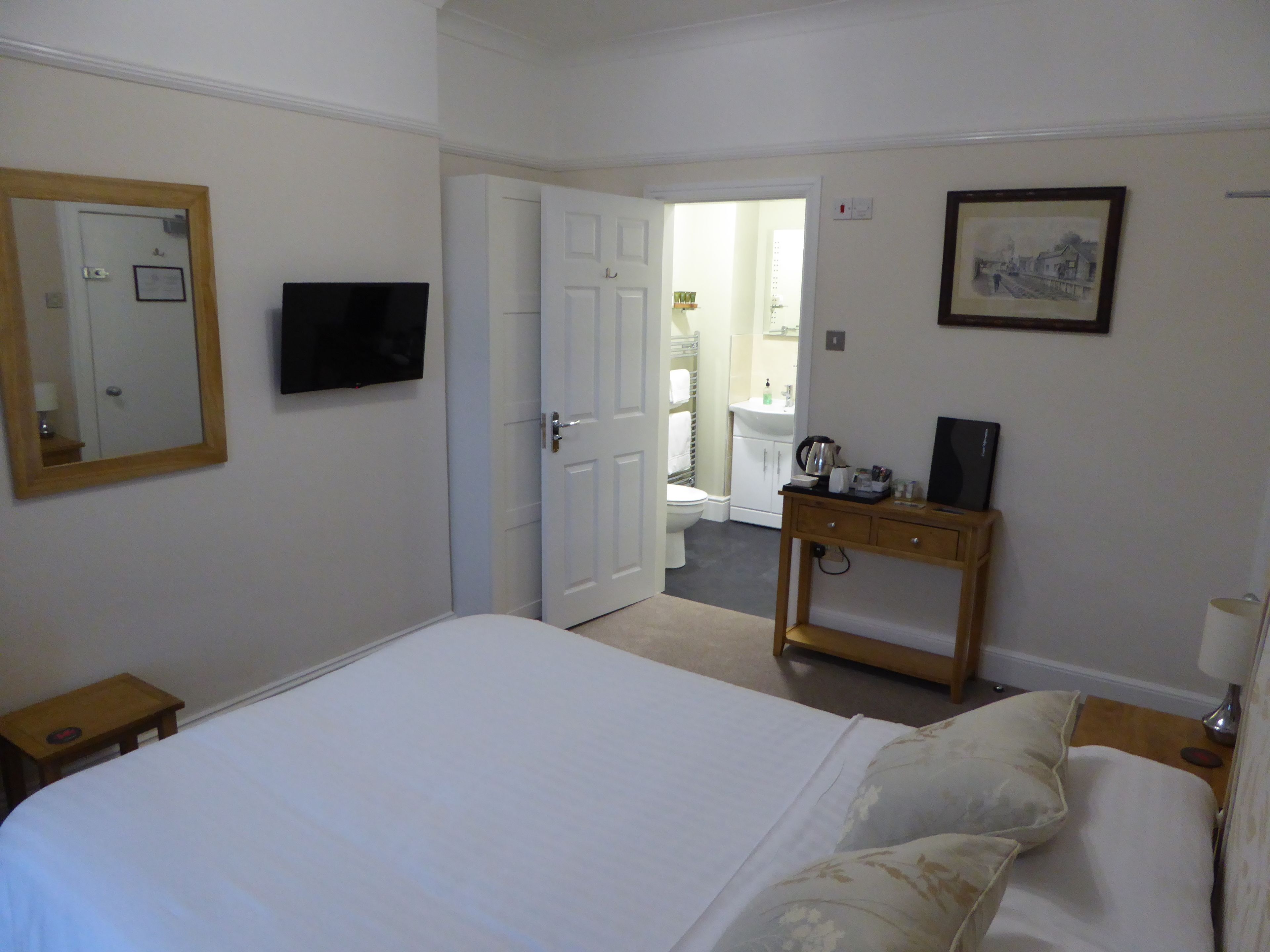 A hotel room with a bed, a bedside table, a TV, a wooden dresser, and a bathroom visible through an open door.