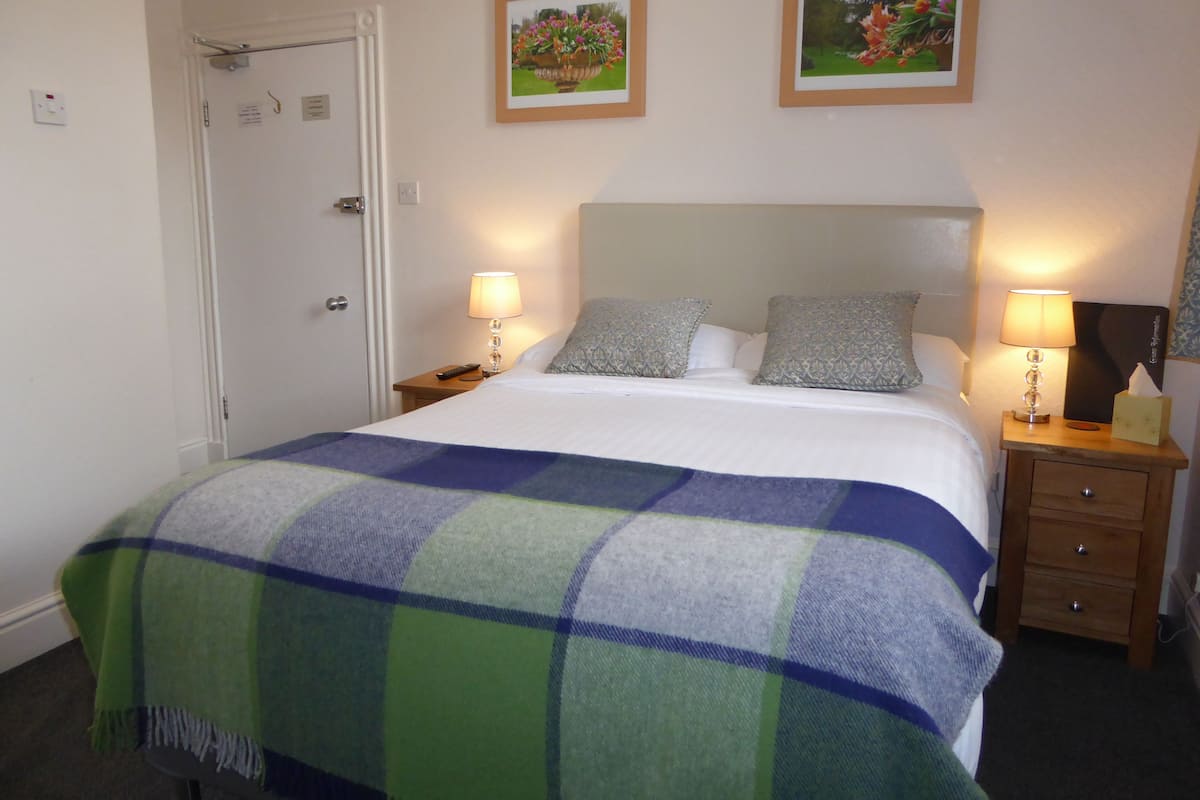 Una cama bien hecha con un edredón escocés, dos lámparas de noche y dos cuadros con flores en la pared.
