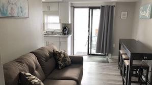 A living room with a brown sofa, a coffee table, and a dining area with a table and chairs. There is a kitchen in the background with white cabinets and a window. - BEST BRIGANTINE VALUE! WE PAY BEACH FEES. WALK TO BEACH, EATERIES. 11AM CHECKOUT (Brigantine)
