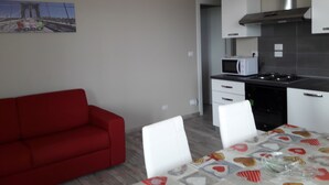 A modern kitchen with white cabinets, a stainless steel range hood, and a microwave. There is a dining table with a colorful tablecloth and white chairs. A red sofa is visible in the foreground. - Villa Sofia Piedmont  (Montaldo Scarampi)
