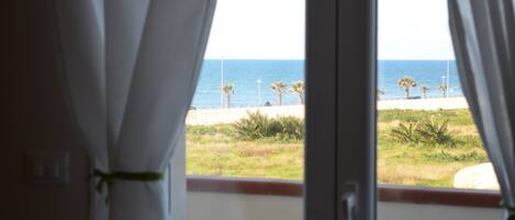 A view through a window with white curtains, featuring a beach with palm trees and a clear sky.
