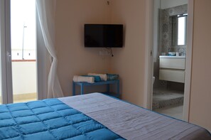 A bedroom with a bed, a TV mounted on the wall, a small table with a towel, and a bathroom visible through an open door.