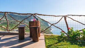 A wooden deck with two cylindrical stools and a table, overlooking a coastal landscape with a clear view of the sea and a distant town. - SUN AND SUN HOUSE (Arco da Calheta)
