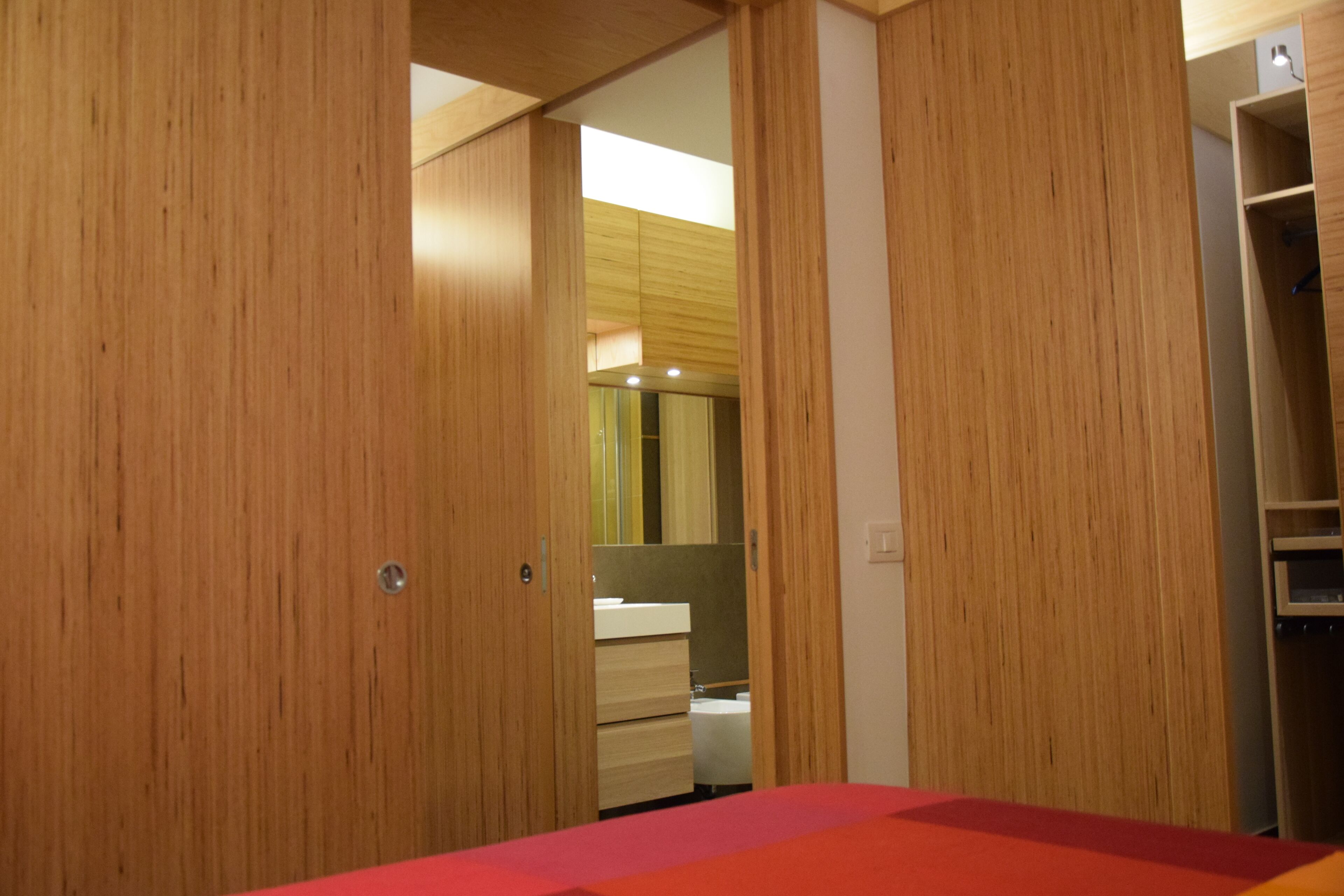 Un dormitorio moderno con armarios de madera, una cama roja y un baño con espejo y lavabo.