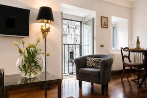 A modern living room with a flat-screen TV, a glass vase with flowers, a dark armchair with a zebra print pillow, and a dining area with a table set for two.