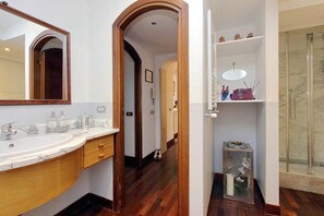 A bathroom with a large mirror, a marble sink, wooden cabinets, a glass shower enclosure, and a hallway leading to another room. - Stunning Pantheon View experience in the heart of Rome (Roma)