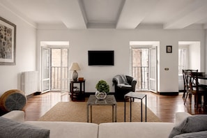 A spacious living room with a sofa, a coffee table, a lamp, and a chair. There is a television mounted on the wall, and a window with a view of the outside.