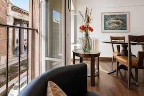 A room with a large window, a dining table with chairs, a vase with flowers, and a painting on the wall.