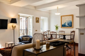 A modern living room with a sofa, a dining table with chairs, a TV mounted on the wall, and a large window.