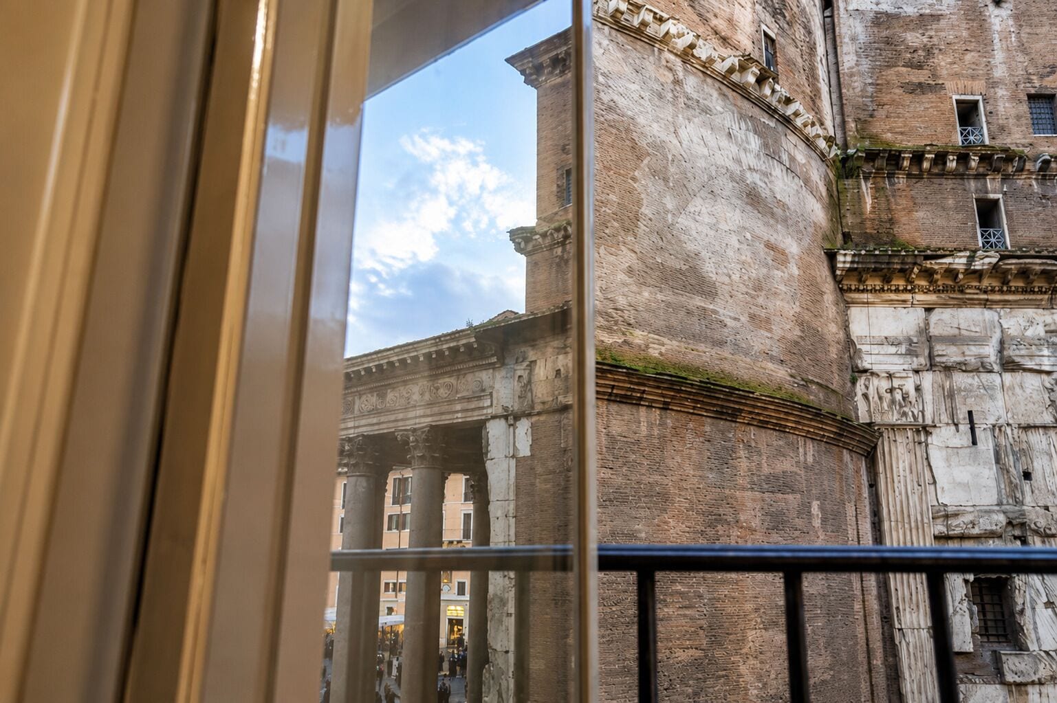 A view through a window with a metal railing, overlooking a historic building with classical architecture, including columns and detailed stonework.