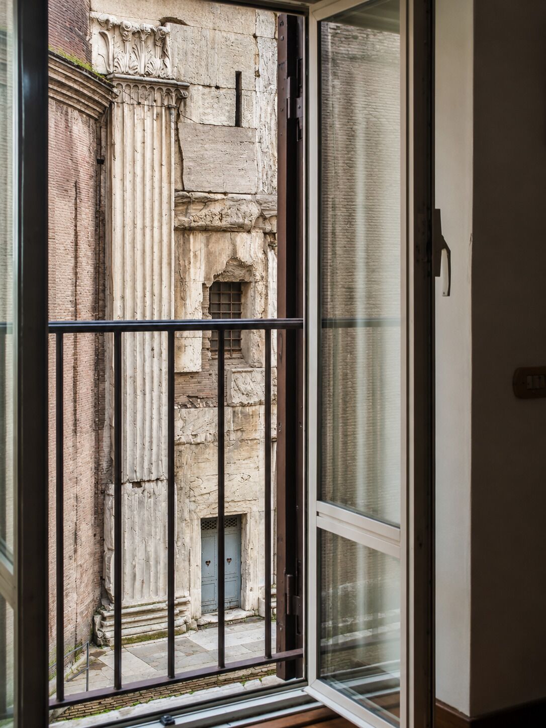 A view through a glass door of a courtyard with a stone wall, a metal gate, and a small window.