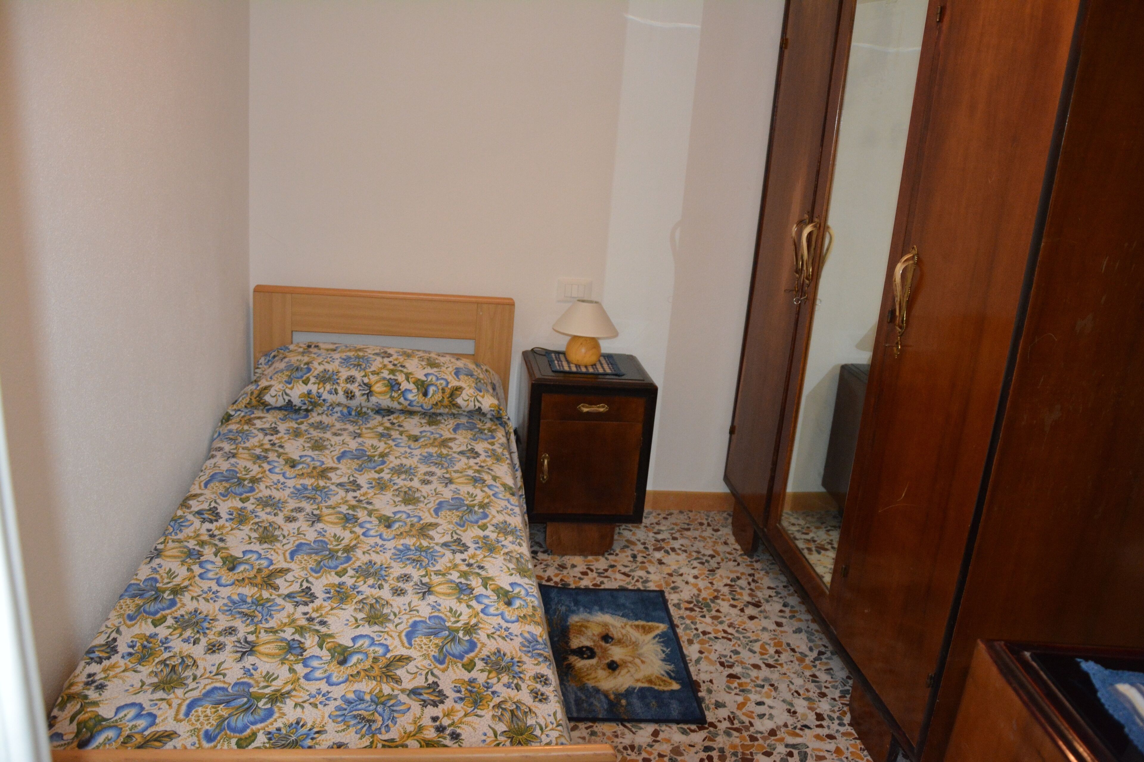 Une petite chambre à coucher avec un lit, une table de chevet, une lampe, une armoire en bois et un animal de compagnie sur un tapis.