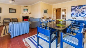 Un salon spacieux avec une table à manger à plateau de verre, entourée de chaises bleues, d’un canapé et d’une cheminée.