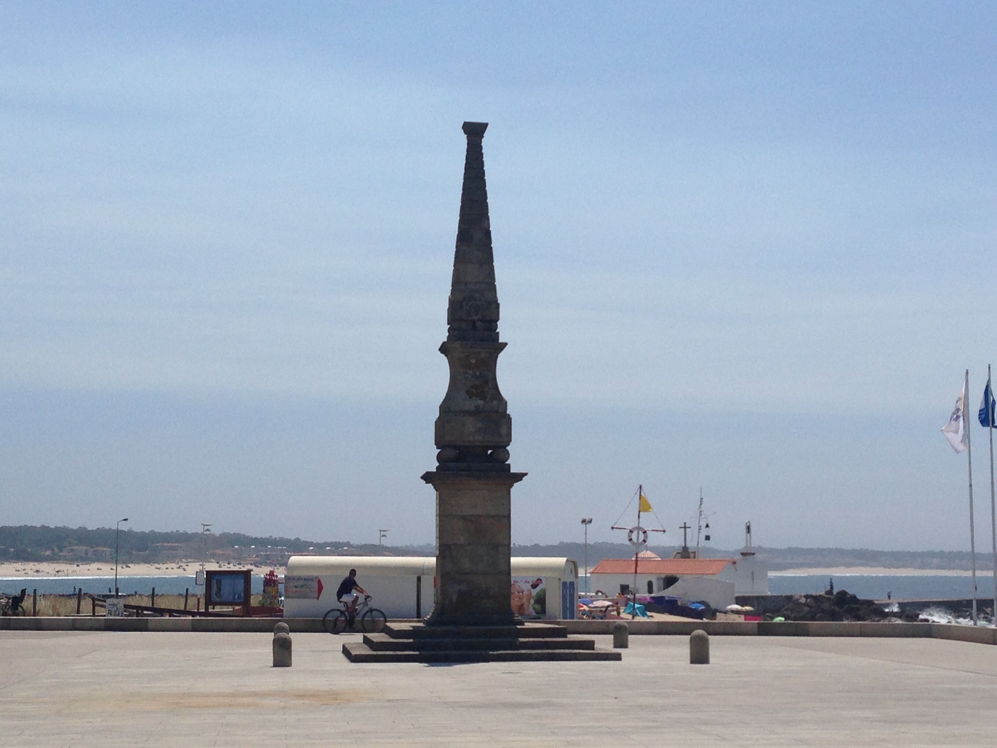 A tall, narrow monument in a plaza with a clear view of the beach and ocean in the background.