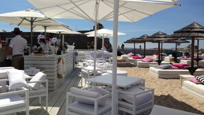 Un espace bar situé au bord de la plage, avec des chaises longues blanches et des parasols, un comptoir de bar et une vue sur la plage et l’océan.