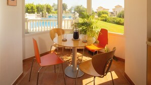 Un comedor iluminado por el sol con mesa y sillas, ventanas amplias con vistas a una piscina y follaje, y una lámpara de techo.
