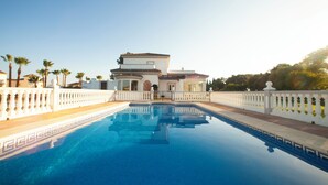 Una piscina grande y rectangular con superficie de agua azul cristalina, rodeada por una valla blanca con barandillas. Al fondo, se observa un edificio amplio de color blanco con tejado de tejas y numerosas ventanas.