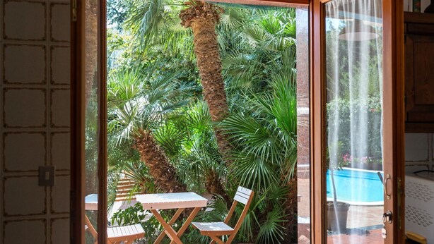 Puerta de vidrio que da a un patio con mesa y sillas, rodeado de exuberante follaje y una piscina visible.