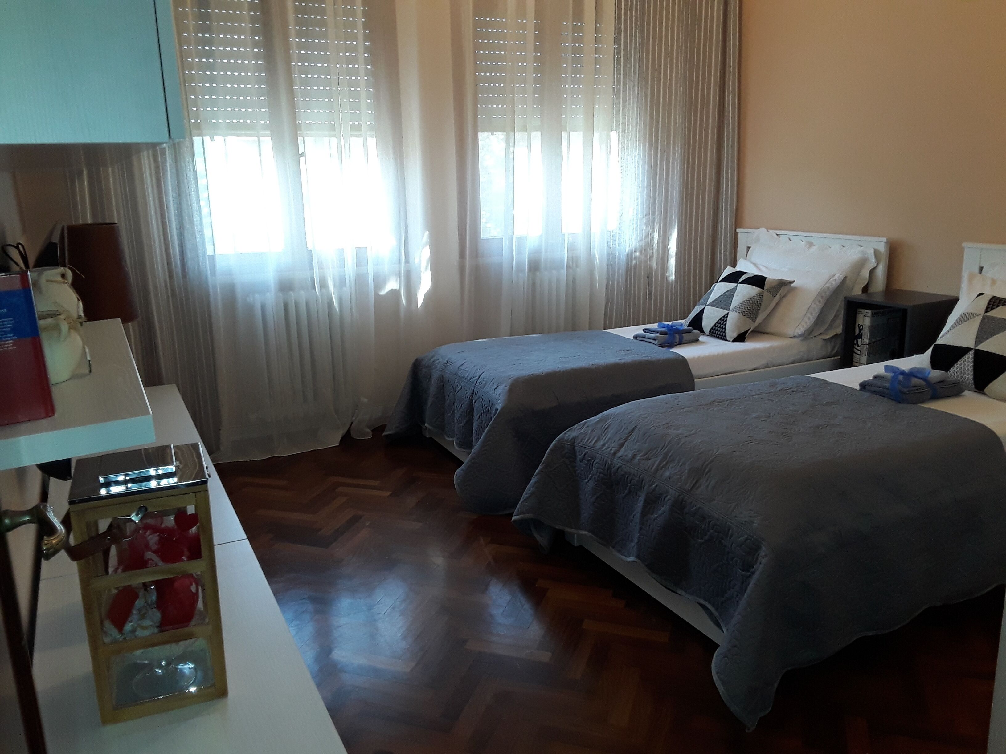 A hotel room with two beds, a wooden floor, a nightstand, and a window with sheer curtains.