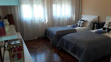 A hotel room with two beds, a wooden floor, a nightstand, and a window with sheer curtains.